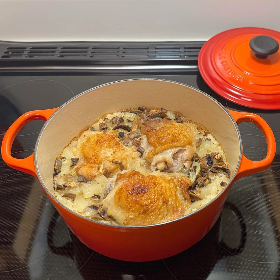 A red Dutch oven filled with golden baked chicken thighs, fluffy long-grain rice, and sautéed mushrooms resting on a glass-top stove.