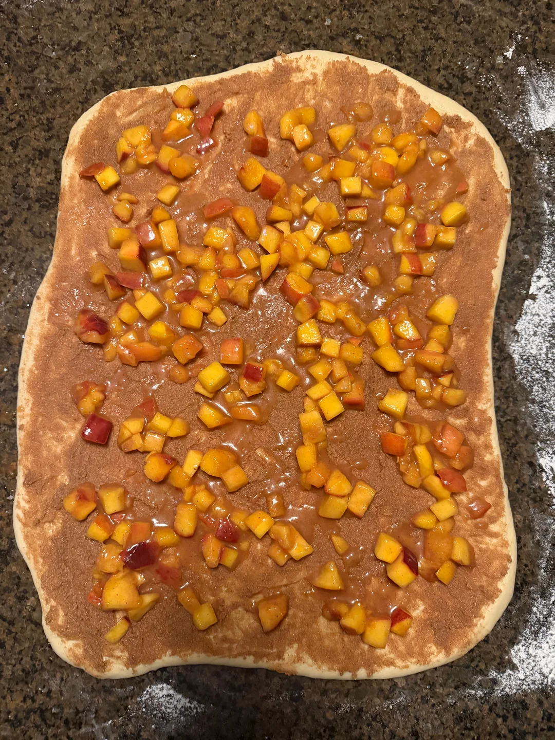 An 18x14-inch rectangle of cinnamon roll dough spread with cinnamon butter and studded with diced peach compote pieces.