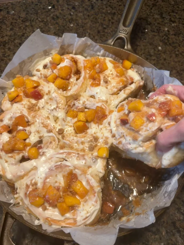 A hand pulling a gooey, frosted sourdough discard peach cobbler cinnamon roll from a parchment-lined 12-inch cast-iron skillet.
