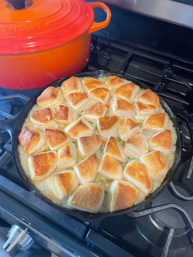 A full cast iron skillet filled with creamy chicken pot pie filling and topped with golden brown, flaky pastry pieces, resting on a black stovetop.