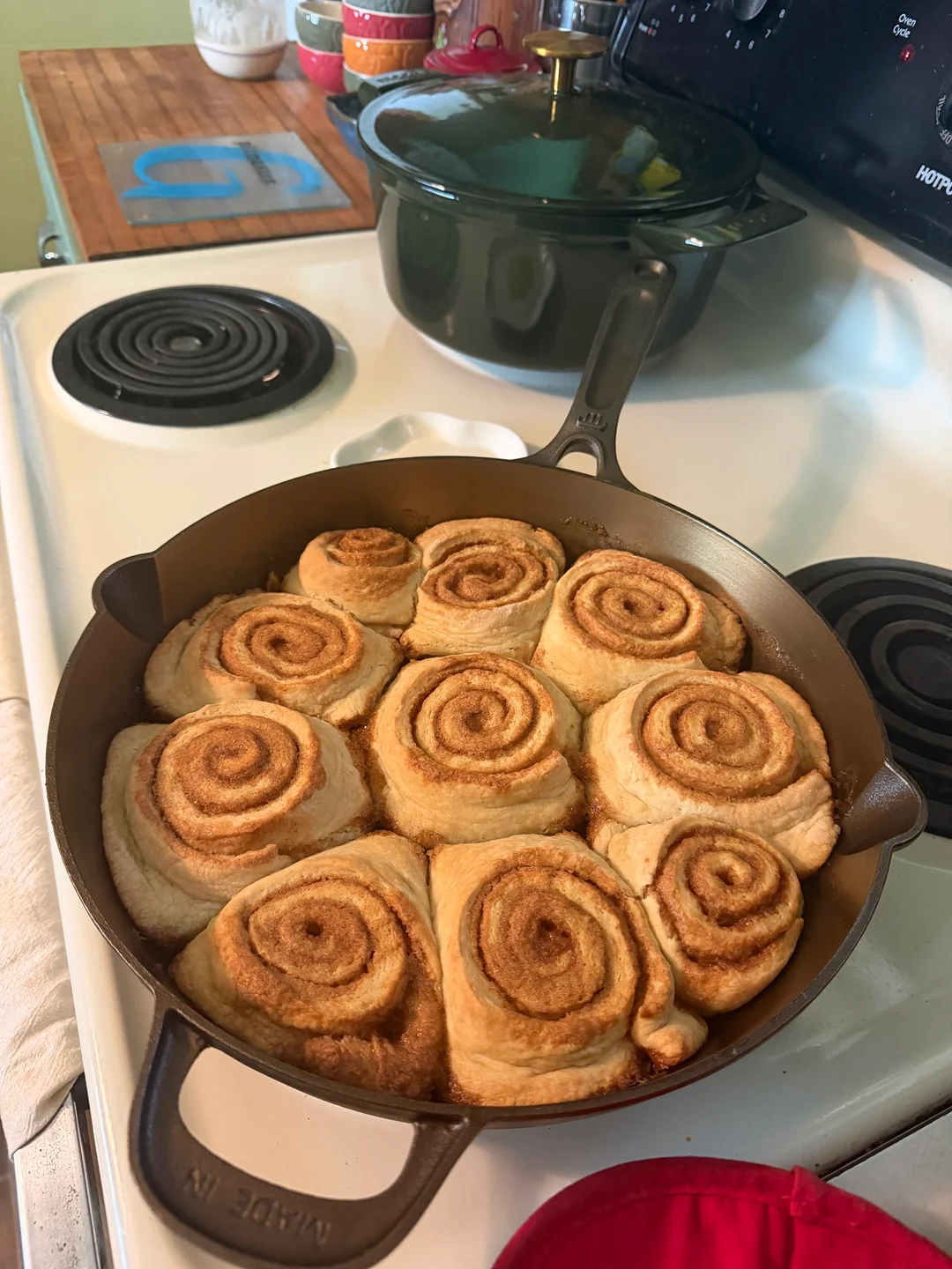 Puffed and risen sourdough cinnamon rolls baked to a golden brown inside a 12-inch cast-iron skillet on a stovetop.