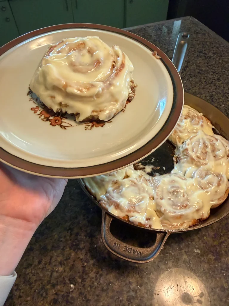 A large plate holding a single frosted sourdough discard cinnamon roll lifted above a 12-inch cast-iron skillet filled with the rest of the baked rolls.