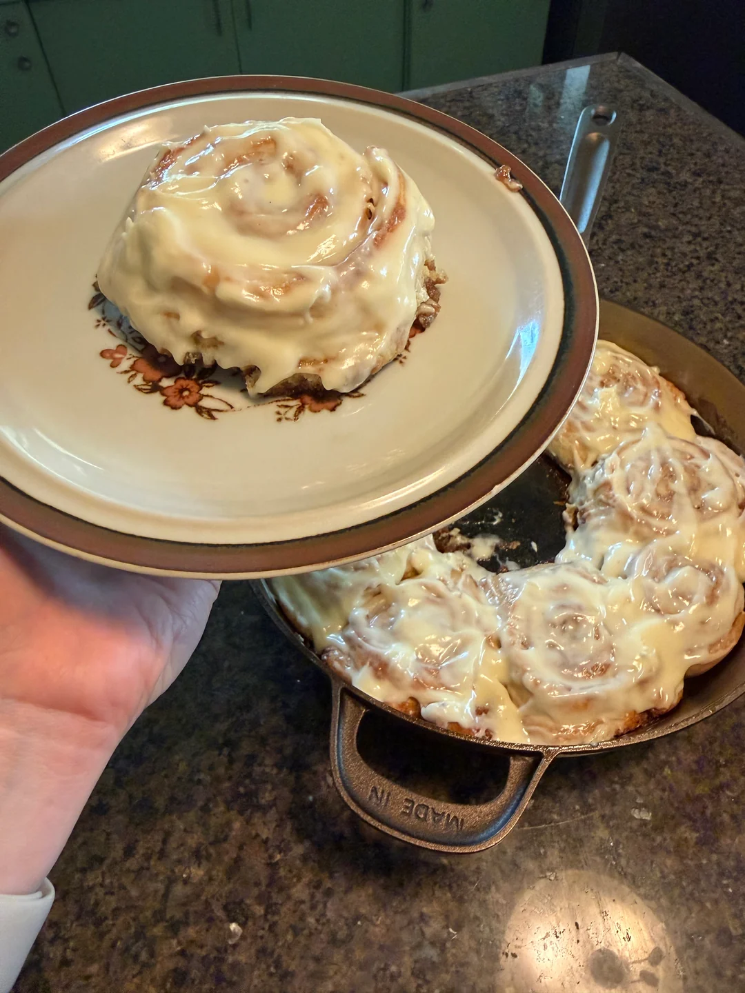 A large plate holding a single frosted sourdough discard cinnamon roll lifted above a 12-inch cast-iron skillet filled with the rest of the baked rolls.