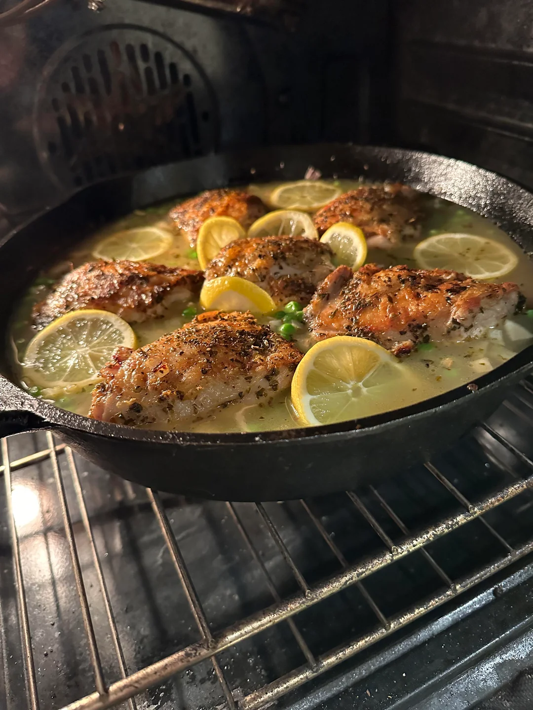 A heavy black cast-iron skillet sitting inside a warm oven, filled with golden-brown chicken thighs baking over a bubbling rice and broth mixture studded with fresh lemon slices and peas.
