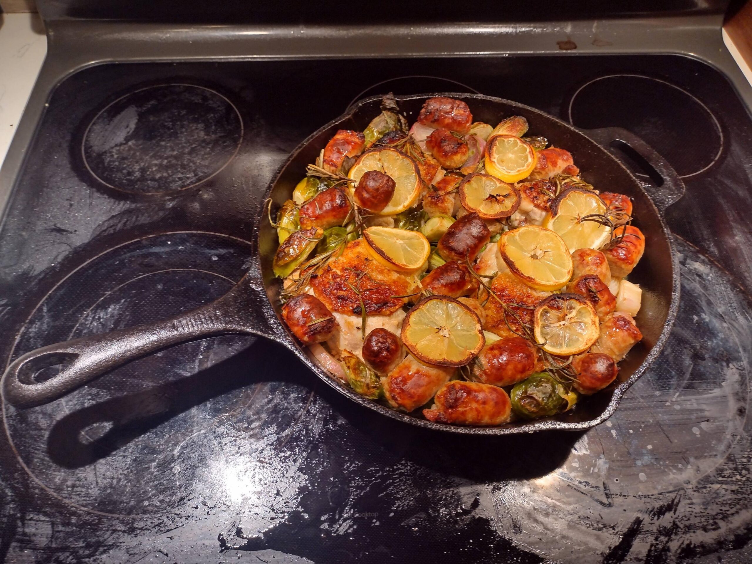 A 12-inch cast iron skillet filled with golden-brown roasted chicken thighs, sliced Italian sausage, caramelized Brussels sprouts, and roasted lemon slices.