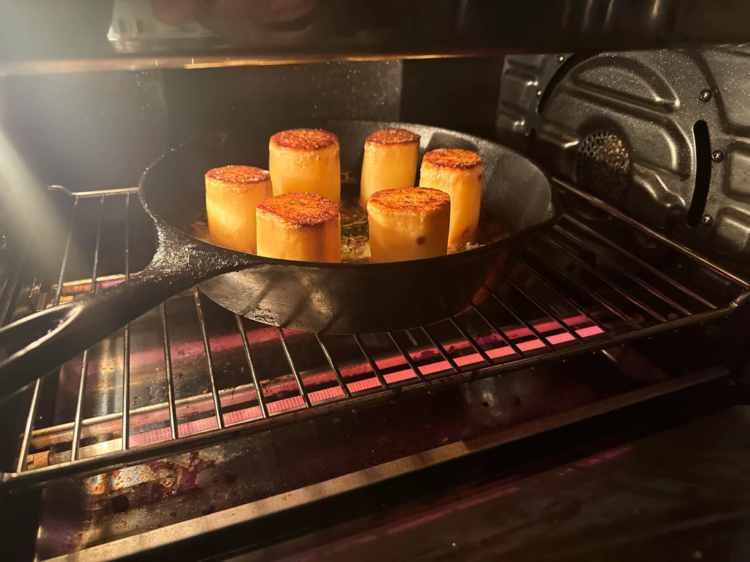 Ten potato cylinders searing in a cast-iron skillet on a stovetop in a pool of bubbling butter and fresh thyme sprigs.